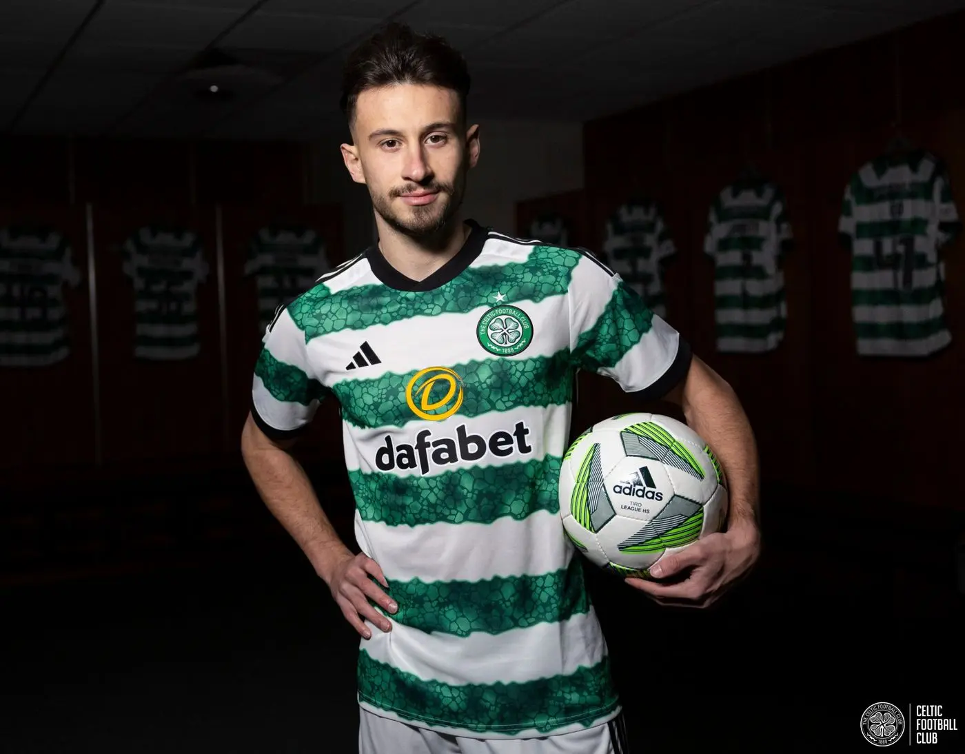 Nicolas Kuhn wearing a Celtic FC jersey, holding a soccer ball in a locker room.