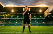 Football referee in black uniform with headset standing authoritatively on stadium pitch under dramatic lighting