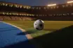 Floodlit football pitch at dusk with corner flag view, blue-white marking lines, and bunting-draped stands in evening light.
