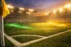 Floodlit football pitch at dusk with emerald and gold lighting, cinematic shadows, corner-flag perspective.