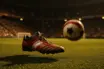 Match ball in motion with football boot on floodlit pitch at evening kickoff, shallow focus with stadium backdrop.