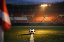 Floodlit stadium corner view with red-and-white seats, football on midfield line under golden spotlight.