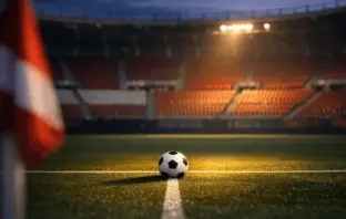 Floodlit stadium corner view with red-and-white seats, football on midfield line under golden spotlight.