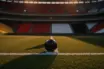 Floodlit football pitch at dusk with match ball on centre spot, dramatic shadows, empty stadium stands in background.
