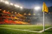 Empty floodlit stadium bowl with red and gold accent lighting, corner flag in foreground, evening atmosphere.