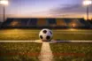 Empty floodlit football pitch at golden hour with ball on penalty spot, dramatic shadows, red-white and blue-gold colour tones.