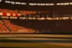 Floodlit international football stadium at golden hour with packed stands, red-white and maroon-white bunting, dramatic pitch lighting, no players.