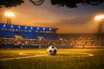 Floodlit football pitch at dusk with ball on centre spot, blue and white stadium lighting, low corner angle.
