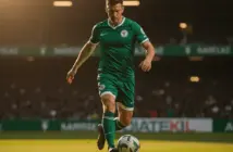 Celtic midfielder in green kit dribbling football on pitch under stadium lights
