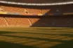 Floodlit stadium pitch at dusk with packed stands and dramatic shadow patterns, golden-hour lighting.