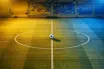 Aerial view of floodlit football pitch with ball on centre spot, golden-yellow and blue stadium lighting, empty stands in soft focus.