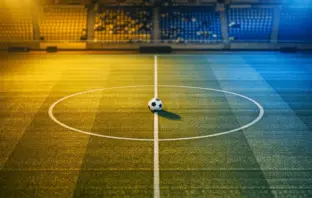 Aerial view of floodlit football pitch with ball on centre spot, golden-yellow and blue stadium lighting, empty stands in soft focus.
