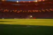 Empty floodlit stadium pitch at dusk with packed stands, dramatic shadows, and amber-blue stadium lighting creating cinematic atmosphere.