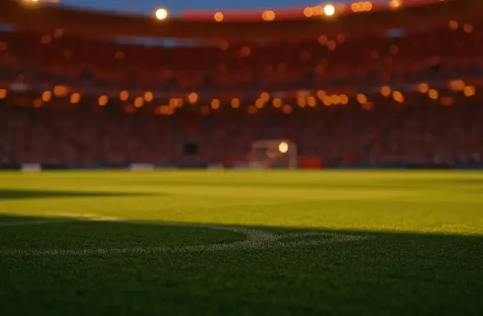 Empty floodlit stadium pitch at dusk with packed stands, dramatic shadows, and amber-blue stadium lighting creating cinematic atmosphere.