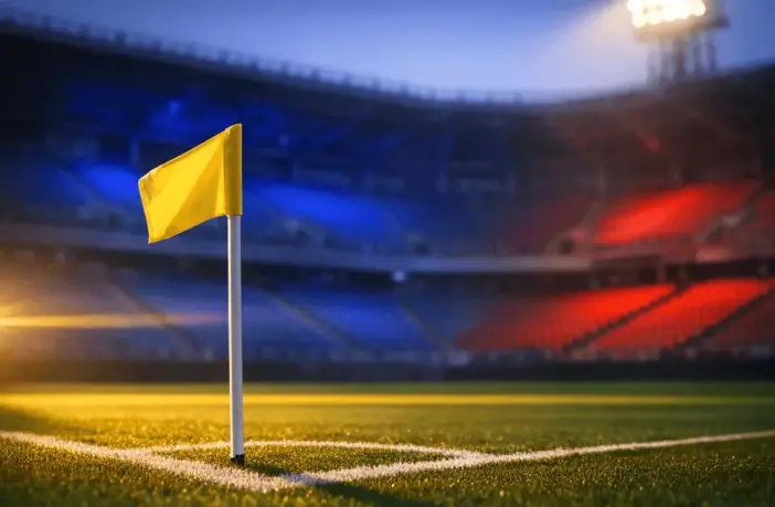 Empty stadium corner with blue-and-red flag-coloured floodlighting, corner flag sharp in foreground, golden dusk light across tiered seating.
