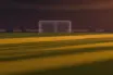 Floodlit football stadium pitch at dusk with packed stands and dramatic shadow patterns, no players visible.
