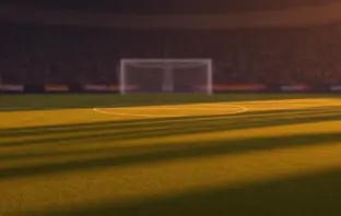 Floodlit football stadium pitch at dusk with packed stands and dramatic shadow patterns, no players visible.