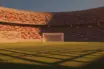 Floodlit football stadium at dusk with goal post shadows and packed stands in national colours, no players