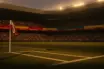 Floodlit football stadium at sunset with packed stands, empty pitch, and gold-hour atmospheric lighting