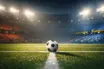 Floodlit football pitch at night with match ball on centre spot, dramatic shadows, empty stadium stands lit in team colours.