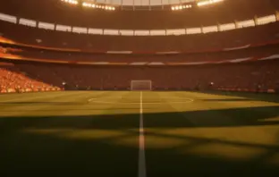 Floodlit international football stadium at sunset with packed stands and shadow patterns across the pitch. No players visible.