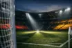 Floodlit stadium lower bowl with German flag-colour lighting over empty pitch at dusk, corner flag perspective.