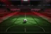 Aerial stadium view of floodlit pitch with match ball on centre circle, red-green colour accent lighting, evening kick-off atmosphere.