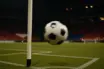 Stadium corner with match ball in motion under floodlights, colour-graded red, blue, white and black.