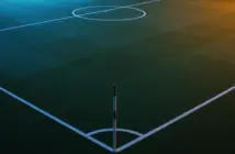 Floodlit football pitch at dusk with geometric markings and dramatic stadium shadows, anticipatory pre-match atmosphere.