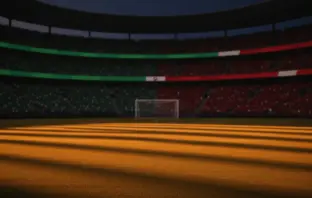 Floodlit football stadium at dusk with green and red crowd sections, dramatic shadow patterns on empty pitch.
