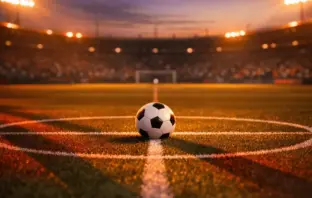 Floodlit football pitch at dusk with ball on centre spot, dramatic shadows, golden stadium lighting, packed stands blurred in background.