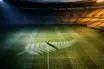 Floodlit stadium pitch at dusk with silver-fern motif and football on centre spot; navy and gold lighting; tournament-ready atmosphere.