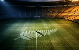 Floodlit stadium pitch at dusk with silver-fern motif and football on centre spot; navy and gold lighting; tournament-ready atmosphere.