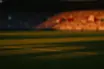 Floodlit stadium pitch at golden-hour kick-off with dramatic shadows and crowd bokeh.
