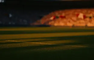 Floodlit stadium pitch at golden-hour kick-off with dramatic shadows and crowd bokeh.
