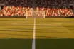 Floodlit football stadium at golden-hour with packed stands, empty pitch, red-white and gold-green crowd colours.