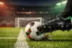 Football boot mid-strike on floodlit pitch with goal net and stadium stands in soft focus during evening kickoff.