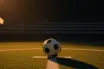 Empty floodlit football pitch with match ball on centre spot, green and blue colour accents, dramatic stadium lighting at dusk.
