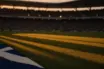 Empty floodlit stadium pitch with blue and yellow shadow patterns and packed stands at dusk