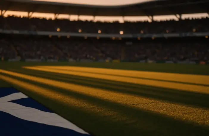 Empty floodlit stadium pitch with blue and yellow shadow patterns and packed stands at dusk
