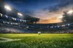 Empty floodlit stadium lower bowl at dusk with blue and white bunting, pitch visible from corner flag angle, match ball on centre spot.