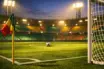Floodlit stadium corner view with green and gold accent lighting, empty pitch, goal net, cinematic sports photography.