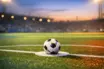 Empty floodlit football pitch at sunset with match ball on penalty spot, blue and red ambient light, dramatic shadows, low-angle shot.