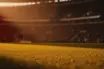 Floodlit football stadium pitch at golden hour with dramatic shadows and packed stands, warm cinematic lighting
