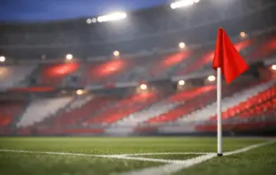 Empty stadium corner with red and white atmospheric lighting, floodlit pitch markings in soft focus, evening mood.