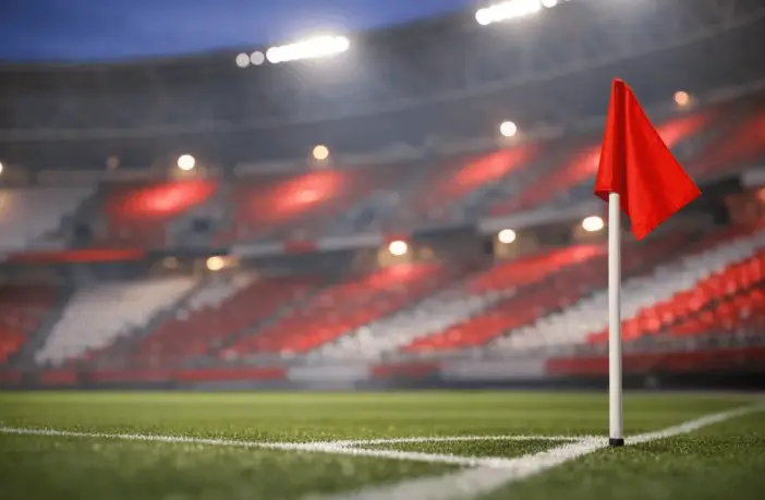 Empty stadium corner with red and white atmospheric lighting, floodlit pitch markings in soft focus, evening mood.