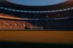 Empty floodlit stadium pitch at dusk with packed stands and dramatic shadows, shot from ground level during golden hour.