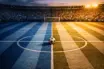 Aerial floodlit stadium view with sky-blue and white pitch markings, golden-hour lighting, and a football on centre spot.
