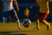 Evening stadium action: players in white-blue and yellow-red kits converging on ball during floodlit match, sharp shadows, shallow focus.