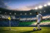Floodlit stadium corner section with blue and green-lit stands; lone player in white kit with ball near corner flag.